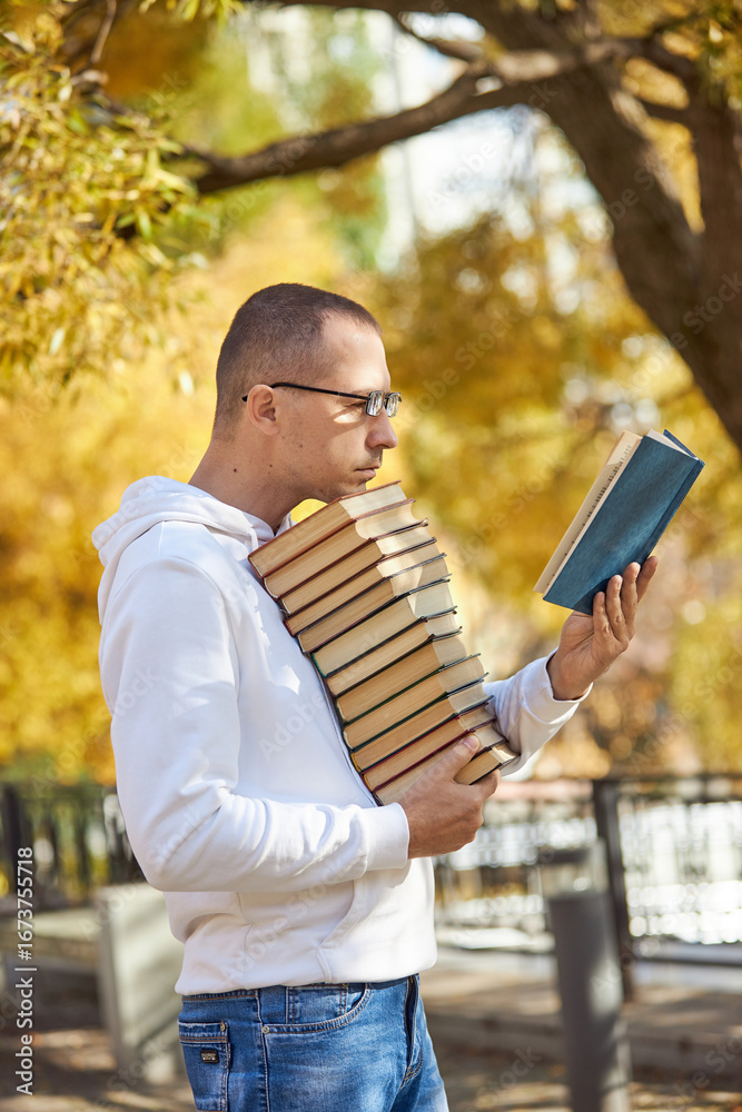 Fototapeta premium A young man stands in a park, enjoying a sunny afternoon. He is holding a book in one hand while balancing a tall stack of books in the other. Gorgeous autumn leaves surround him