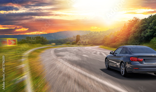 A sleek black car drives quickly along a curved road at sunset with motion blur, surrounded by trees, hills, and a glowing sky in a scenic landscape.