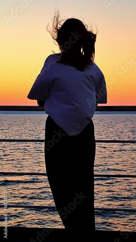 A silhouette of a young woman with their hair blowing in the wind, watching the sunset over the sea from a ship's deck. Vertical video.