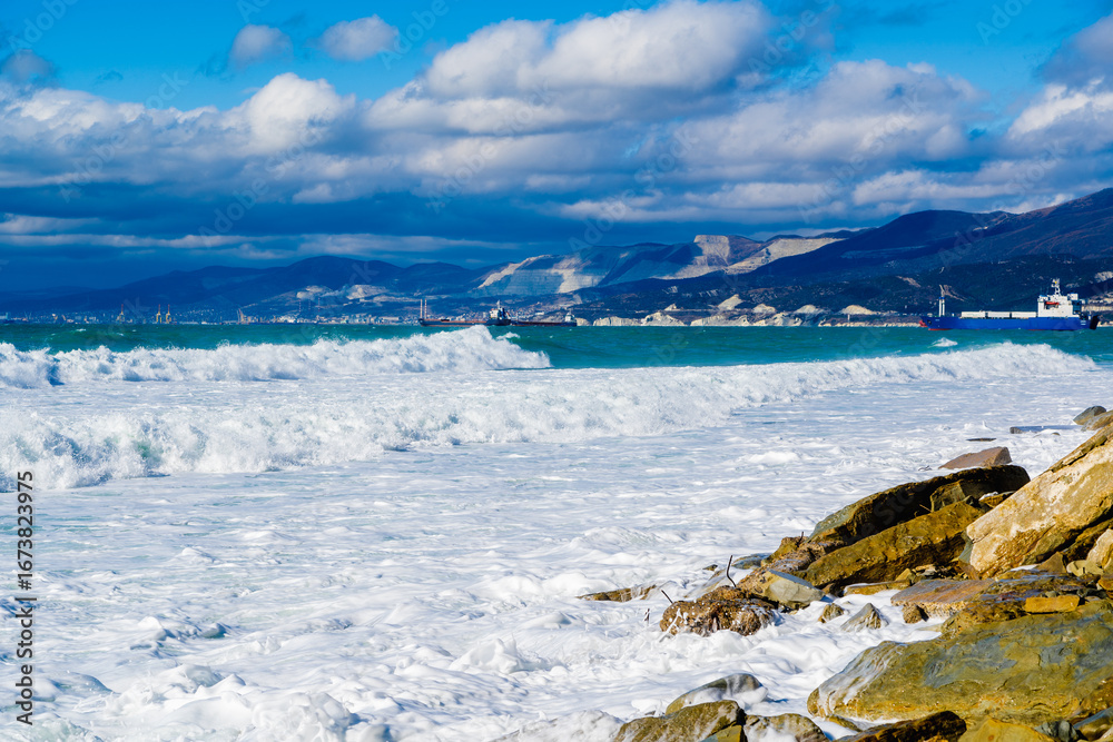 Obraz premium Kabardinka resort. Large green storm waves in white foam roll over rocky beach of Kabardinka. In background is port of Novorossiysk and Caucasus Mountains. Large stones on shore. 
