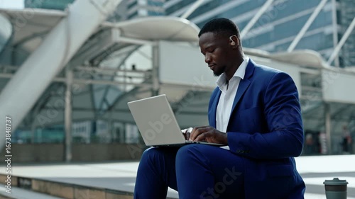 Portrait of African American man with hearing aid works on laptop near office center. Businessman with disability writes report on notebook computer sitting on downtown street