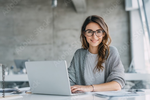 Smiling young woman with glasses working on laptop in modern office environment.