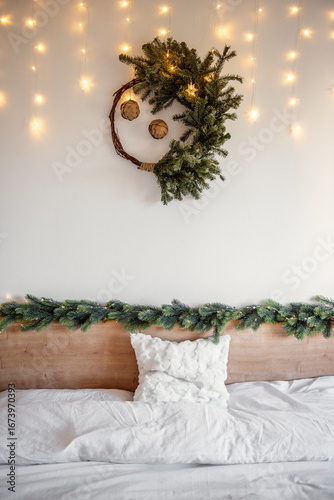 Christmas wreath adorned with pine cones hanging above a bed, with fairy lights softly illuminating a cozy bedroom, creating a warm and festive atmosphere for the holiday season. Festive interior 