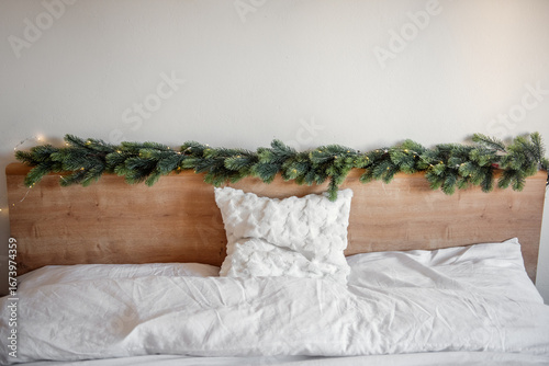 Christmas garland with small lights decorating the wooden headboard of a bed with white sheets and a white pillow, creating a cozy and festive atmosphere