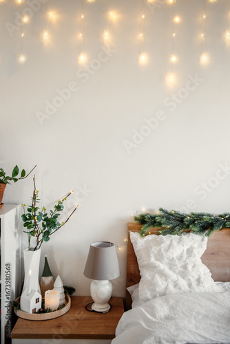Christmas lights are hanging on a white wall and decorating a bed headboard with white pillows and blankets. A bedside table with a lamp. Plants and Christmas decorations. Creating a cozy atmosphere