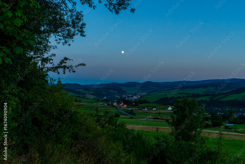 Obraz premium Picturesque rural valley with fields, houses, and winding roads under a rising full moon. The calm twilight atmosphere highlights harmony between village and nature.
