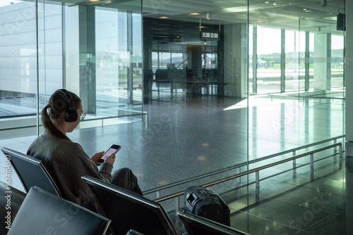Young woman sitting at airport gate using smartphone and looking at her digital boarding pass. Wearing headphones and waiting for flight with luggage beside. Modern travel concept.