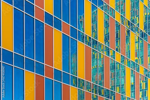 Facade of a modern building with vertical glass panels in blue, yellow, and orange tones. Reflections of a nearby structure add depth and visual complexity.