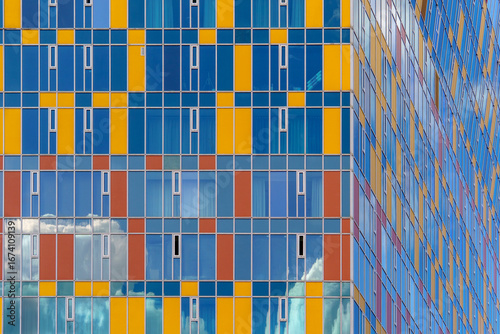 Facade of a modern building featuring a grid of rectangular panels in blue, yellow, and reddish-orange. Glass sections reflect the sky and clouds, adding a dynamic visual effect.