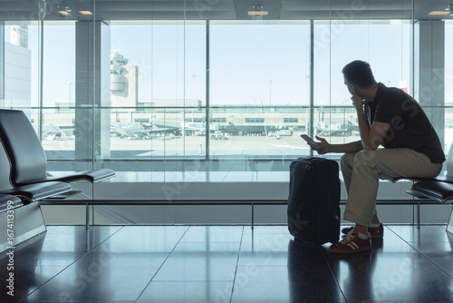 Young man sitting at airport gate waiting for a flight, looking out the window at airplanes with smartphone and suitcase. Modern air travel concept.