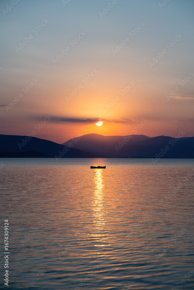 Obraz premium Tranquil fishing boat at sunset over Tolo Bay Greece. Glowing orange sky and distant mountain silhouettes reflecting on calm sea water
