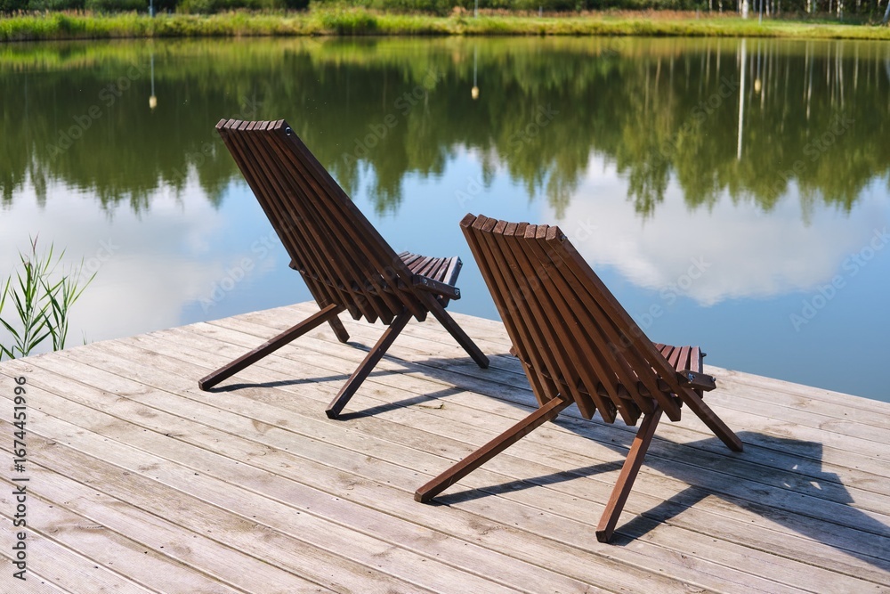 Fototapeta premium Brown wooden sun loungers on the deck on the shore of a forest lake. Rest, relaxation.