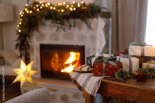 Stylish christmas presents, ornaments, gingerbread cookies on table on background of decorated fireplace with lights in nordic living room in magical eve. Merry Christmas!