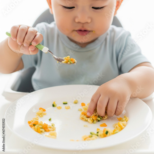 A heartwarming and authentic shot of an adorable Asian baby boy's hands eating food from a plate by himself while sitting in a high chair