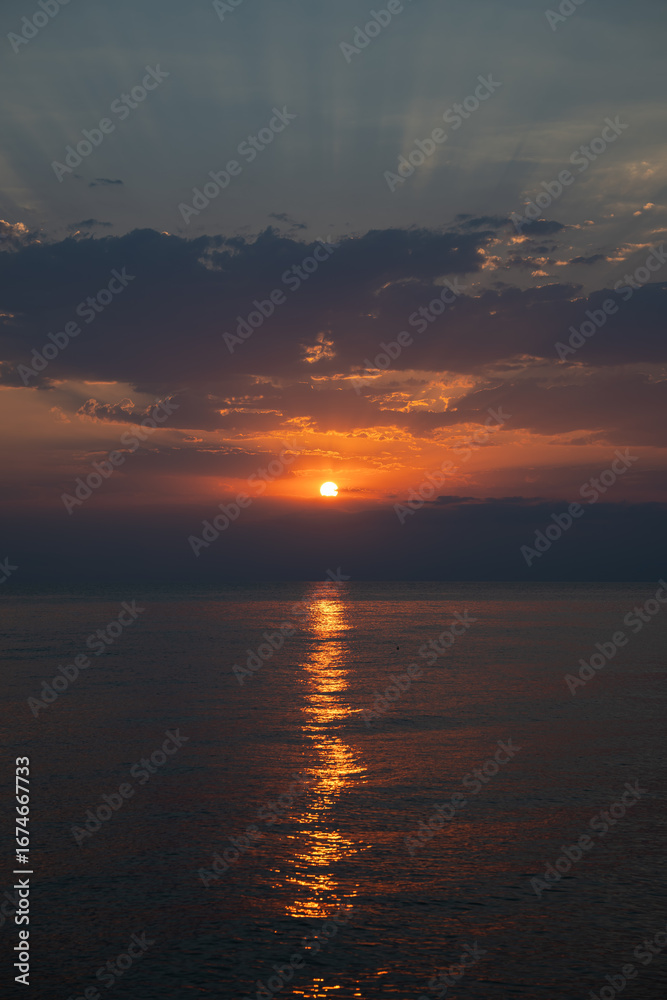 Naklejka premium Dramatic golden sunrise over sea with breaking through dark clouds. Peaceful and serene horizon at dawn as the sun rises and reflects on calm water. Hope and new beginnings