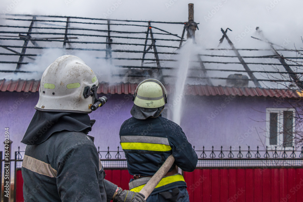 Fototapeta premium Two firefighters spray water on a house's partially collapsed roof, working to extinguish a fire. The image, taken from behind, highlights their teamwork and the ongoing emergency response