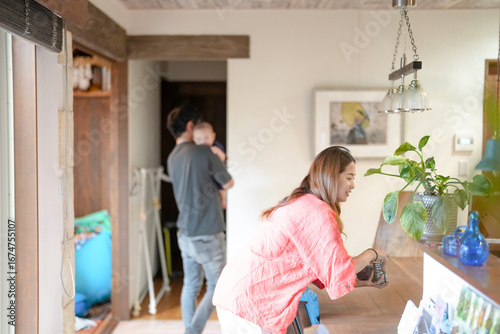Fotografie In a Mediterranean-style living room, a Japanese man in his 30s walks holding a 3-month-old baby while a mother in her 20s wearing orange cleans the table