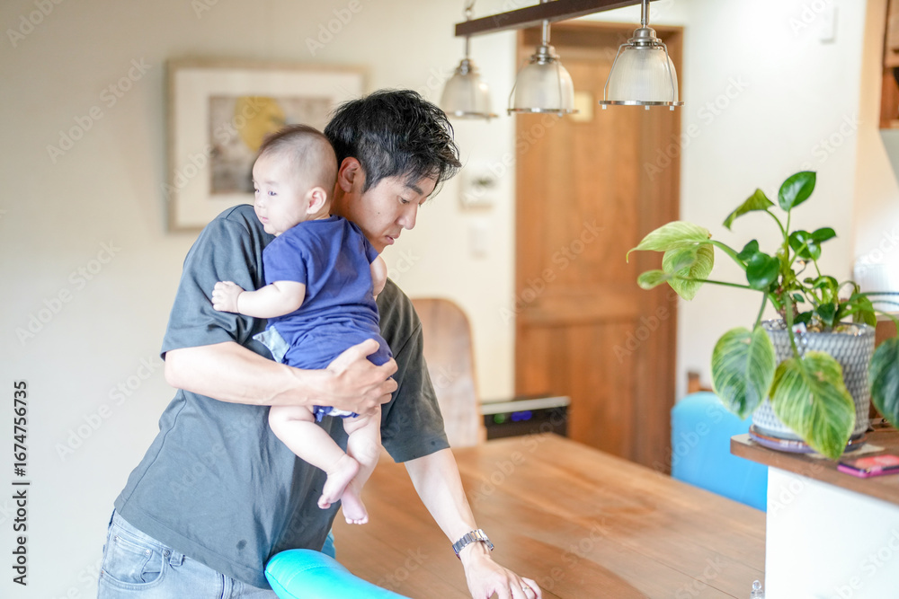 Obraz premium In a Mediterranean-style living room, a Japanese man in his 30s wipes the table with a cloth while holding a 3-month-old baby. Summer. Japan.