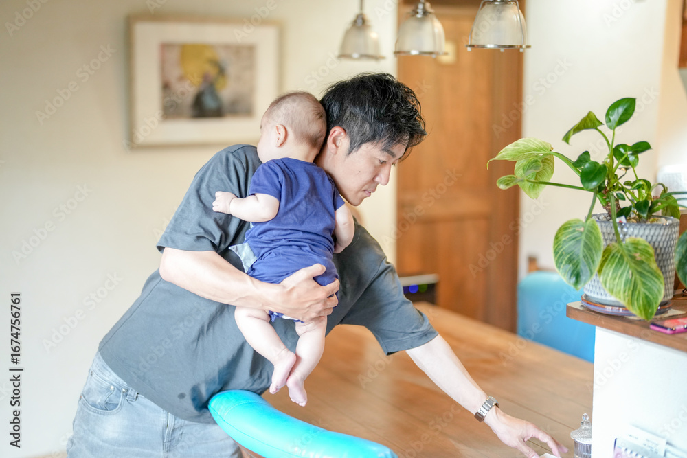 Obraz premium In a Mediterranean-style living room, a Japanese man in his 30s wipes the table with a cloth while holding a 3-month-old baby. Summer. Japan.
