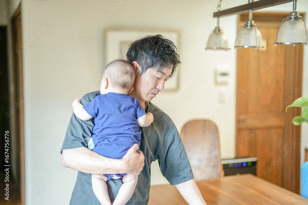 Obraz premium In a Mediterranean-style living room, a Japanese man in his 30s wipes the table with a cloth while holding a 3-month-old baby. Summer. Japan.