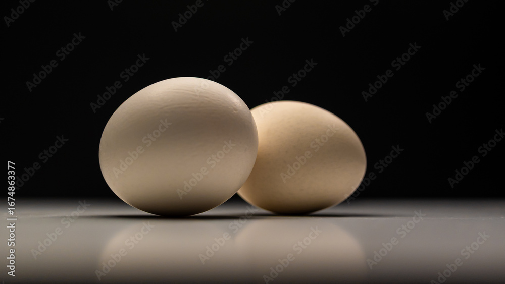 Fototapeta premium Two light brown chicken eggs are positioned on a white surface, casting soft shadows against a dark, blurred background, creating a simple still life.