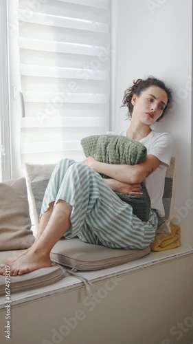 Emotional moment of a woman seated in a cozy living room, embracing a pillow while gazing thoughtfully out the window and looks very lonely and sad