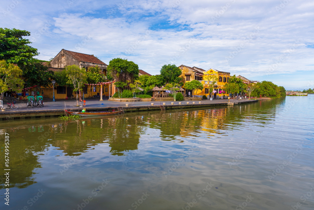 Obraz premium A panoramic view of Hoi An's waterfront, with traditional yellow buildings and vibrant trees reflecting on the calm river, a beautiful scene under a bright, cloudy sky.