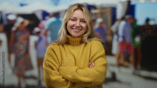Wallpaper Mural Smiling woman with long blonde hair and yellow sweater stands with arms crossed in a lively outdoor market on a sunny city day. Torontodigital.ca