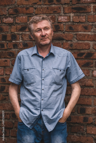 authentic portrait middle-aged man standing casually against old brick wall, hands in his pockets, wearing blue checkered shirt. he appears pensive, reflecting urban life around him. close up.