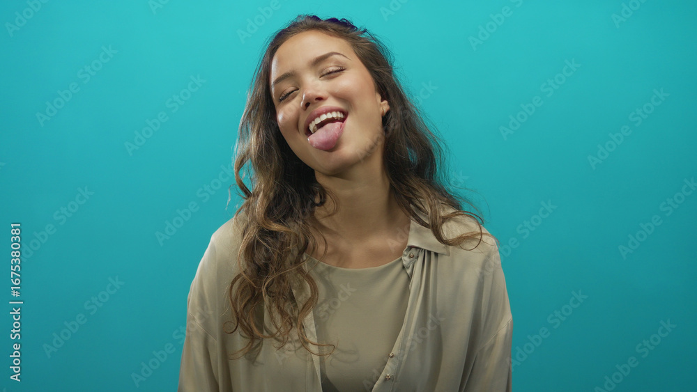 Obraz premium Woman sticks out tongue with long hair loose over blue studio wall, wearing beige blouse; playfulness.