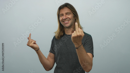 Blond man with long hair shows middle finger gesture with both hands in studio under light blue wall; rebellion.
