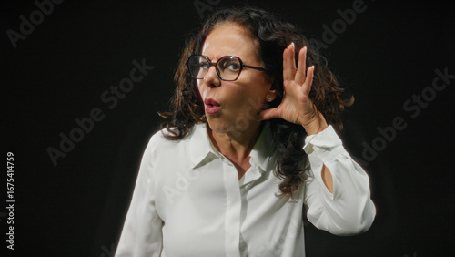 Woman with curly hair and glasses showing surprise on black background, wearing white shirt and expressing emotion through hand gesture while isolated in darkness.