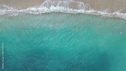 Drone shot of turquoise ocean waves breaking on sandy shore, creating natural abstract seascape with foam and texture.