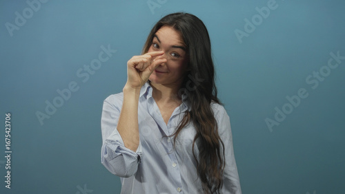 Young hispanic woman pinching fingers to show small size while smiling in blue studio setting; playfulness.