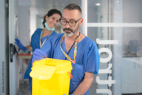 Doctor disposing of needle in sharps container with nurse in background