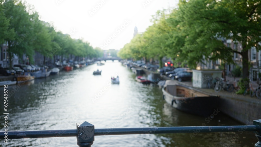 Naklejka premium Blurred canal scene in amsterdam with boats floating on water, trees lining the banks, and defocused cityscape in the background creating a serene dutch atmosphere.