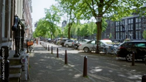 Amsterdam cityscape with blurred street view of parked cars, bicycles, and leafy trees along a picturesque urban canal on a sunny day with shadows on the walkway.