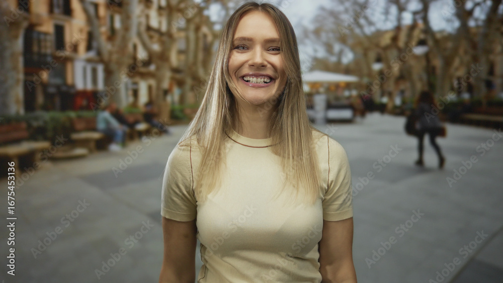Fototapeta premium Young blonde woman smiling happily outdoors on a vibrant city street with blurred background, exuding energy and joy in an urban setting.