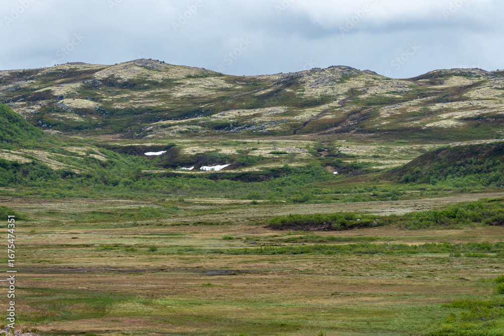 Obraz premium Rolling hills adorned with patches of snow contrast with vibrant green vegetation. The overcast sky adds a mystical quality to the tranquil landscape, creating a peaceful atmosphere.