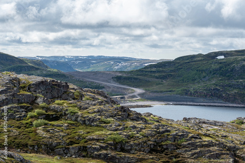 A wide landscape reveals rocky terrain and a serene body of water nestled among rolling hills. Clouds float overhead, adding drama to the serene atmosphere in a remote northern area.