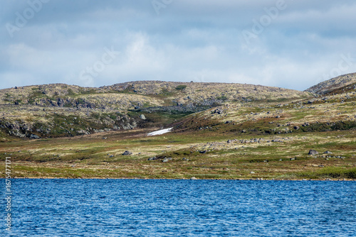 Rolling hills covered in green vegetation and scattered boulders surround a calm blue lake under a cloudy sky. The peaceful landscape showcases nature's untouched beauty.