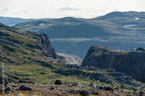 A vehicle travels along a winding road through rugged hills and rocky landscapes, set against a backdrop of distant mountains in soft evening light. The scene captures nature's raw beauty.