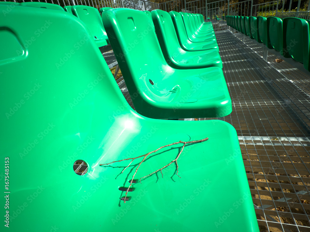 Obraz premium Closeup of green plastic stadium seats on outdoor bleachers with small dry twig lying on chair, empty audience seating in park.