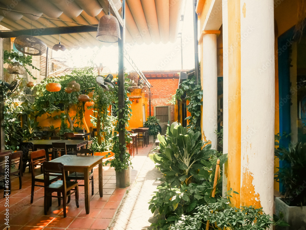 Naklejka premium Warm, sun-kissed yellow iconic wall of narrow alleys converted heritage homes in Hoi An cafe with red-tiled floor, hanging plants, rattan lights. Wooden tables, garden-style dining space