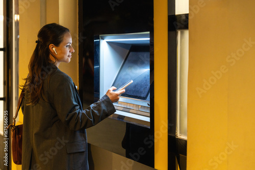 Obraz na plátně Woman using smartphone for contactless ATM cash withdrawal on city street at nig
