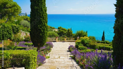 A walkway with a view of the ocean and a garden with purple flowers. The walkway is made of stone and has a railing