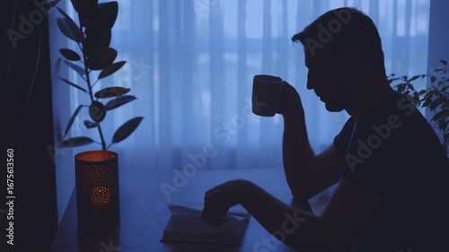 dark silhouette man close up hand flip through paper pages enjoy reading book near window dusk blue light home room candle. guy sitting at desk hand holding cup tea coffee hot drink. hobby rest relax