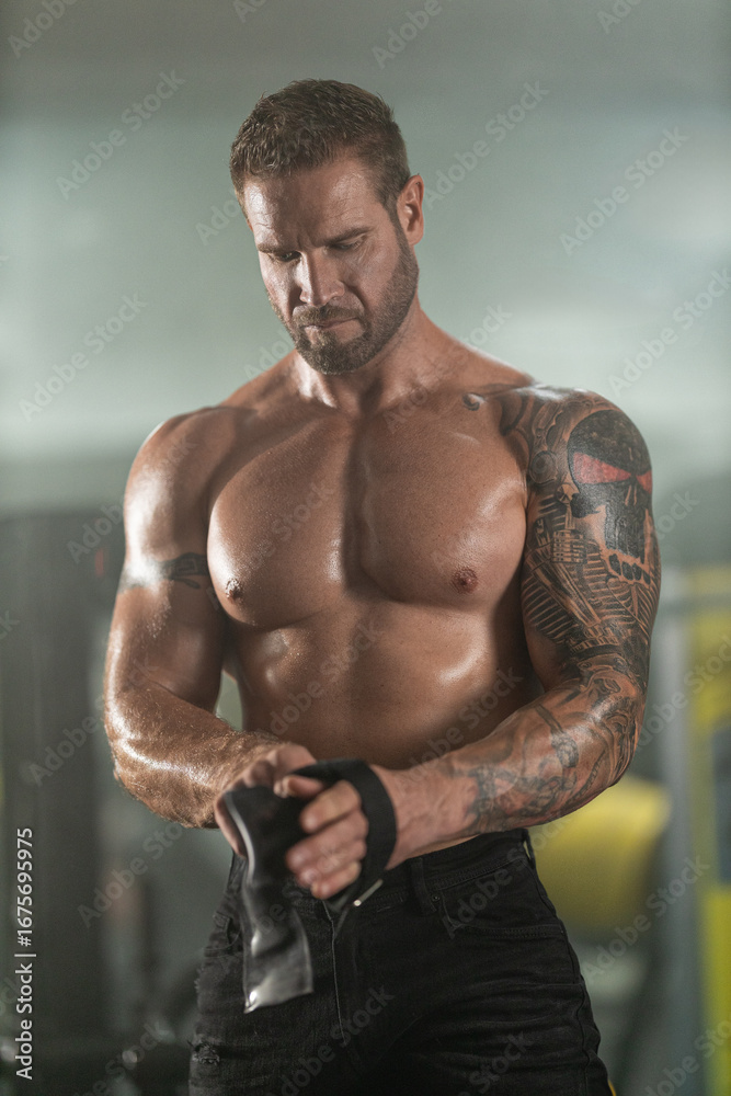 Fototapeta premium muscular man stands in a gym tightening straps on his arm in preparation for a weightlifting workout. The atmosphere is filled with fitness equipment and a focus on strength training.