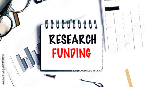 Research funding phrase on notebook with charts, pen, glasses and calculator on white office desk