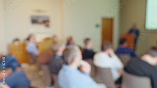 Defocused video of a speaker presenting to an audience in a meeting room. Blurred background with screen graphics, perfect as an active backdrop for titles.